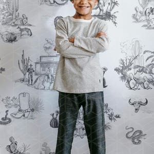 May include: A young person wearing a striped long-sleeve shirt, gray patterned pants, and a gray beanie. The background features a desert-themed wallpaper with cacti, cowboy boots, and other western illustrations.