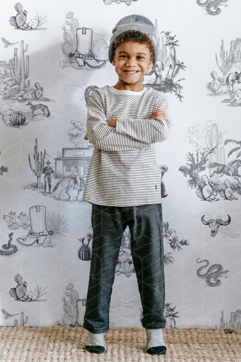 May include: A young person wearing a striped long-sleeve shirt, gray patterned pants, and a gray beanie. The background features a desert-themed wallpaper with cacti, cowboy boots, and other western illustrations.