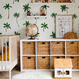 May include: A white crib with a yellow blanket and a white fluffy rug in front of a white dresser with wicker baskets. The wall is covered in a white and green palm tree wallpaper. There is a globe, a wooden toy house, and a framed alphabet chart with animal illustrations.