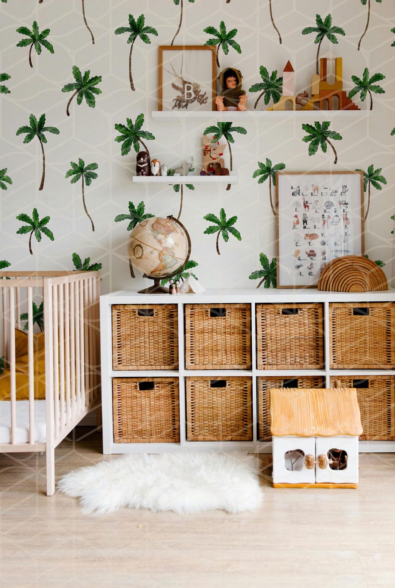 May include: A white crib with a yellow blanket and a white fluffy rug in front of a white dresser with wicker baskets. The wall is covered in a white and green palm tree wallpaper. There is a globe, a wooden toy house, and a framed alphabet chart with animal illustrations.