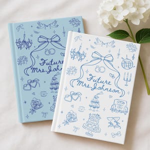 May include: Two wedding journals, one pale blue and one white, with blue illustrations. The covers feature a bow, rings, and the text "Future Mrs. Johnson". Decorative elements include a cake, champagne glasses, and fireworks.