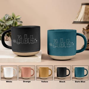 May include: Two ceramic mugs, one black and one teal, with a light beige base and handle. Each mug features a white line drawing of three cats with the names "Pepper", "Fluffy", and "Bella". Additional mugs in white, orange, yellow, black, and dark blue are shown.