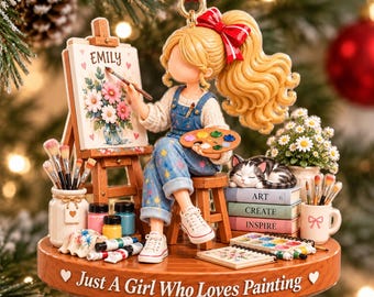 以下が含まれることがあります： 「Emily」と書かれたイーゼルで絵を描く金髪の人物、画材、猫、Art、Create、Inspireと書かれた本が配置された装飾品。台座には「Just A Girl Who Loves Painting」と刻まれています。