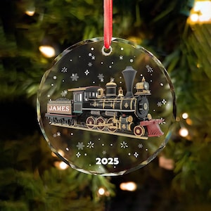 May include: A clear, round Christmas ornament with a detailed black and gold train design. The train is labeled "JAMES" and the year "2025" is displayed. The ornament is decorated with snowflakes and star shapes, and hangs from a red ribbon.