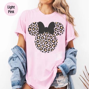 Peut inclure: T-shirt rose clair avec un n&oelig;ud noir et un motif t&ecirc;te de souris l&eacute;opard. Le texte "Light Pink" est dans un cercle rose en haut &agrave; gauche. La chemise est port&eacute;e avec une veste en jean.