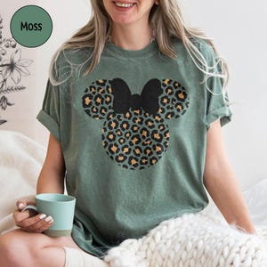Peut inclure: T-shirt vert mousse avec un motif Mickey Mouse &agrave; imprim&eacute; l&eacute;opard et un n&oelig;ud noir. Le t-shirt a des manches courtes et une coupe d&eacute;contract&eacute;e. Le mot "Moss" est dans un cercle vert en haut &agrave; gauche.