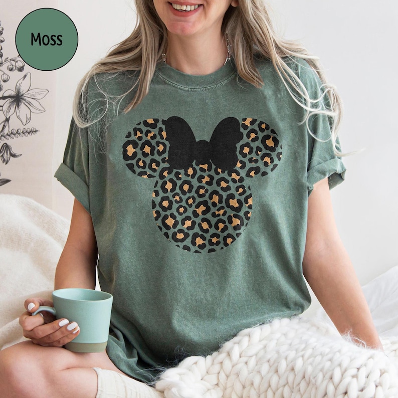 Peut inclure: T-shirt vert mousse avec un motif Mickey Mouse &agrave; imprim&eacute; l&eacute;opard et un n&oelig;ud noir. Le t-shirt a des manches courtes et une coupe d&eacute;contract&eacute;e. Le mot "Moss" est dans un cercle vert en haut &agrave; gauche.