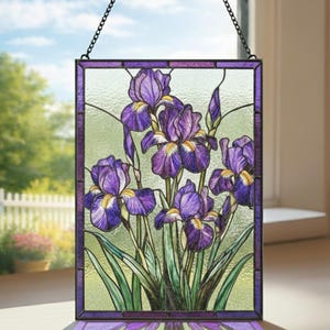 May include: Stained glass panel featuring purple iris flowers with yellow centers and green leaves. The artwork is framed in a dark purple border and hangs from a chain. The background is a light green color.