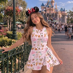 May include: A sleeveless romper featuring a light pink background with an all-over print of various snacks, including ice cream cones, donuts, and popcorn buckets, alongside mouse-ear shaped balloons. The garment has a wrap-style skirt overlay.