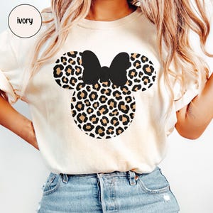 Peut inclure: T-shirt ivoire avec un motif Minnie Mouse. Le motif comprend un n&oelig;ud noir et des oreilles et un visage &agrave; imprim&eacute; l&eacute;opard. Le mot "ivoire" est dans un cercle en haut &agrave; gauche.