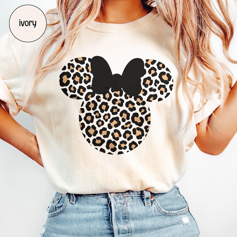 Peut inclure: T-shirt ivoire avec un motif Minnie Mouse. Le motif comprend un n&oelig;ud noir et des oreilles et un visage &agrave; imprim&eacute; l&eacute;opard. Le mot "ivoire" est dans un cercle en haut &agrave; gauche.
