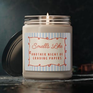 May include: A lit candle in a glass jar with a black lid. The label reads "Smells Like Another Night of Grading Papers" in red script and a blue and white striped background. The candle is on a dark surface.