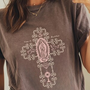 May include: A dark gray t-shirt featuring a pink cross design with religious imagery and the words "Virgen de Guadalupe" in a decorative font. The shirt has rolled-up sleeves and a relaxed fit.