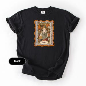 May include: Black t-shirt featuring a framed Aries zodiac sign design. The design includes a ram's head surrounded by flowers, with the word "Aries" at the bottom. The shirt is made of a soft material.