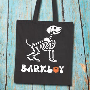 May include: Black tote bag featuring a white dog skeleton design. The word "BARKLEY" is printed in white, with an orange ghost graphic. The bag has black handles and is made of a durable fabric.