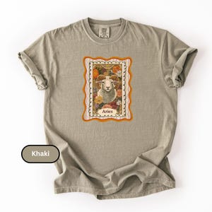 May include: Khaki-colored t-shirt featuring a framed Aries zodiac sign graphic. The graphic includes a ram's head surrounded by flowers, with the word "Aries" at the bottom. The shirt has a classic crew neck and short sleeves.