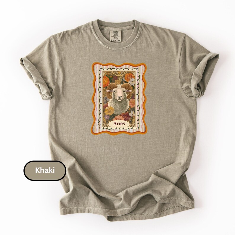 May include: Khaki-colored t-shirt featuring a framed Aries zodiac sign graphic. The graphic includes a ram's head surrounded by flowers, with the word "Aries" at the bottom. The shirt has a classic crew neck and short sleeves.