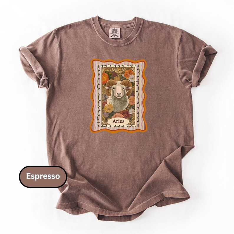 May include: Espresso-colored t-shirt featuring a framed Aries zodiac sign graphic. The graphic includes a ram surrounded by flowers and the word "Aries". The shirt has a crew neck and short sleeves.