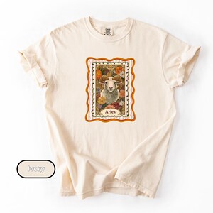May include: Ivory-colored t-shirt featuring a framed Aries zodiac sign graphic. The graphic includes a ram's head surrounded by flowers, with the word "Aries" below. The shirt has rolled-up sleeves and a crew neck.