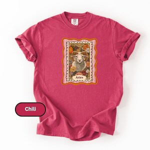May include: A chili-colored t-shirt featuring a framed illustration of a ram with the word "Aries" below. The ram is surrounded by flowers in a vintage style. The shirt has rolled-up sleeves.