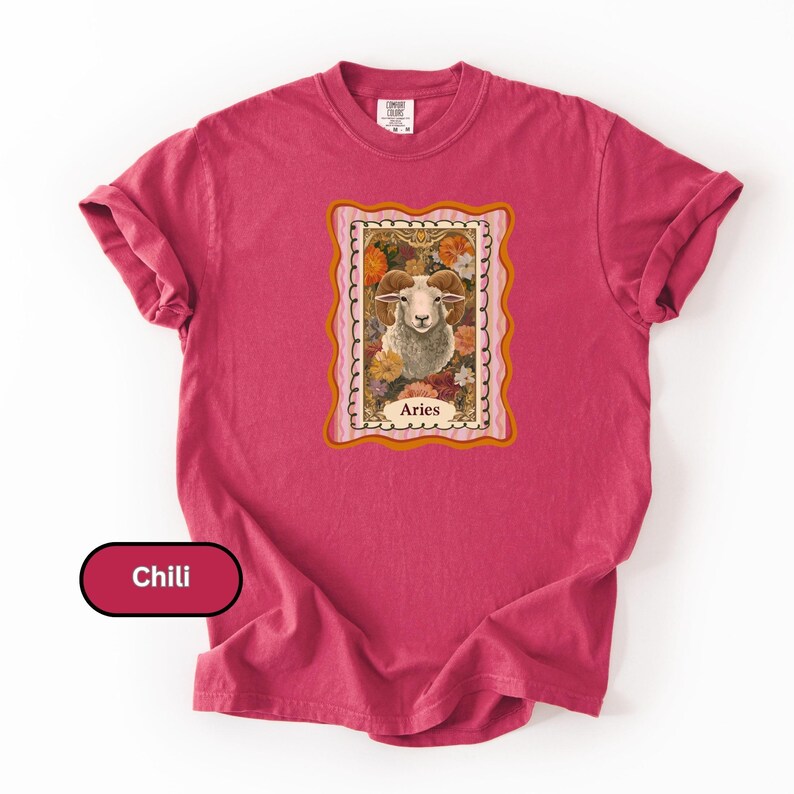 May include: A chili-colored t-shirt featuring a framed illustration of a ram with the word "Aries" below. The ram is surrounded by flowers in a vintage style. The shirt has rolled-up sleeves.