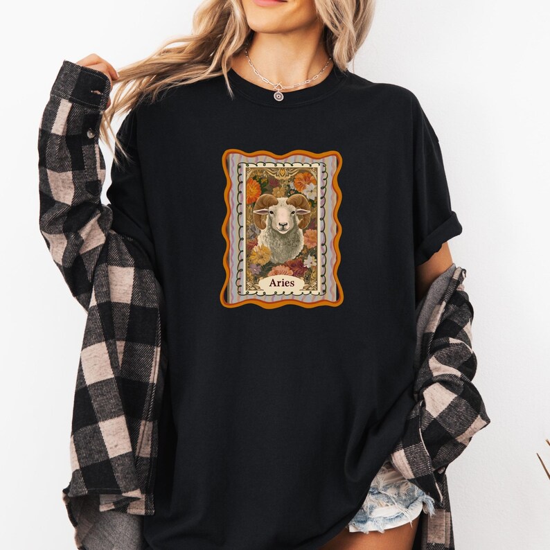 May include: Black long-sleeve shirt featuring a framed illustration of an Aries ram surrounded by flowers. The illustration has a gold border and the word "Aries" at the bottom. The shirt is worn with a black and tan plaid jacket.