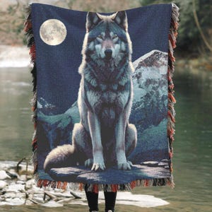 May include: A woven throw blanket with a detailed illustration of a wolf under a full moon. The blanket has a dark blue background with mountain and water imagery. The edges are fringed with red, grey, and black threads.