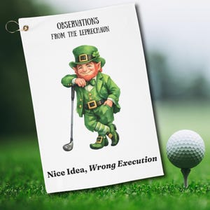 May include: White golf towel with a cartoon leprechaun in a green suit, leaning on a golf club. Text reads "OBSERVATIONS FROM THE LEPRECHAUN" and "Nice Idea, Wrong Execution". A golf ball sits on a tee in the green grass.
