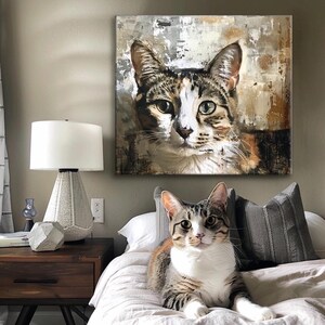 May include: A tabby cat with white markings rests on a bed, mirroring a large canvas painting of a similar cat. The painting features a textured background with brown, white, and gray tones. A white lamp and wooden nightstand are in the background.