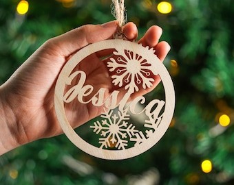 May include: A wooden Christmas bauble with the name "Jessica" cut out in a cursive font. The bauble features snowflake designs and is suspended by a natural twine string. The background is a blurred Christmas tree.