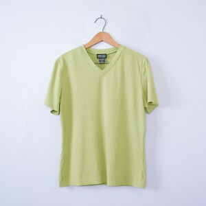 Puede incluir: Una camiseta verde claro con cuello en V y mangas cortas, con textura acanalada vertical, colgada de una percha de madera. La etiqueta en el cuello dice "NY&Co". El fondo es blanco.