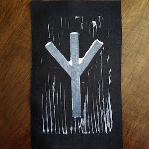 May include: A black rectangular patch featuring a white, stylized rune symbol. The symbol is a vertical line with three lines branching out from the top. The background has vertical white brushstrokes.