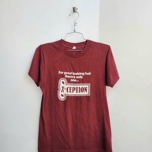 Pode incluir: T-shirt bordô com texto branco: "For good looking hair there's only one... &CEPTION". A t-shirt está pendurada em um cabide de metal contra uma parede branca.