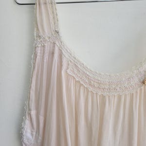 May include: A pale pink vintage nightgown with delicate lace trim and a scoop neckline. The sleeveless garment is made of a lightweight, sheer fabric and hangs from a metal hanger against a white wall. The nightgown has a romantic, feminine aesthetic.