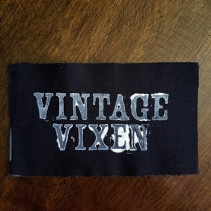 May include: A rectangular black fabric patch with the words "VINTAGE VIXEN" printed in a distressed, light gray font. The patch is set against a dark wooden background, creating a contrast.