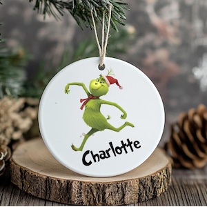 May include: A white, circular Christmas decoration with a green cartoon character in a Santa hat and red scarf, and the name "Charlotte" in black script. It hangs from a natural twine string.