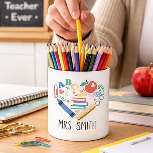May include: A white pencil pot filled with coloured pencils, featuring a heart-shaped design with school-themed illustrations and the text "MRS SMITH". A hand holds a yellow pencil. A chalkboard with "Best Teacher Ever" is in the background.