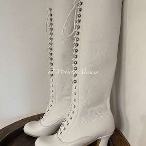 May include: White leather knee-high boots with lace-up fronts and silver eyelets. The boots have a chunky heel and a rounded toe.