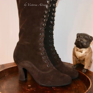 May include: A pair of brown suede lace-up boots with a high heel. The boots are on a wooden tray with a small figurine of a dog wearing a tuxedo.