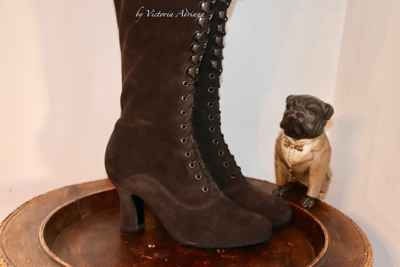 May include: A pair of brown suede lace-up boots with a high heel. The boots are on a wooden tray with a small figurine of a dog wearing a tuxedo.