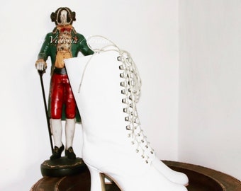 May include: A pair of white, mid-calf boots with a lace-up design. The boots have a small heel and are displayed on a wooden tray. A decorative figurine in a green coat and red pants stands in the background.