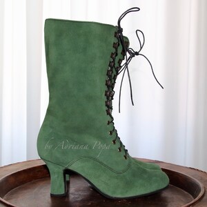 Könnte beinhalten: Grüne Wildleder-Wadenstiefel mit kleinem Absatz und dunkelbraunen Schnürsenkeln. Die Stiefel haben einen Vintage-Stil mit Schnürdesign und sind aus weichem, strukturiertem Material gefertigt. Die Stiefel werden auf einer Holzoberfläche präsentiert.