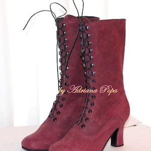 May include: A pair of burgundy-colored, mid-calf boots with a lace-up design. The boots have a small heel and a rounded toe. The laces are black, and the boots appear to be made of suede or a similar material. The text "by Adriana Popa" is visible.