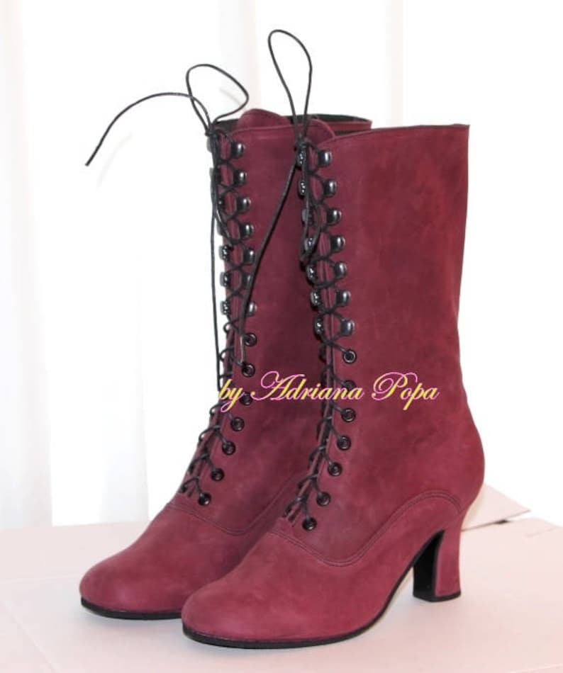 May include: A pair of burgundy-colored, mid-calf boots with a lace-up design. The boots have a small heel and a rounded toe. The laces are black, and the boots appear to be made of suede or a similar material. The text "by Adriana Popa" is visible.