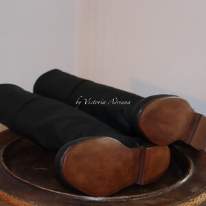 May include: A pair of black leather knee-high boots with wooden soles. The boots are on a wooden surface. "by Victoria Adriana" is written on the boots.