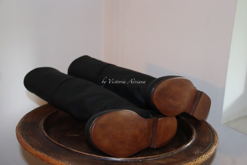 May include: A pair of black leather knee-high boots with wooden soles. The boots are on a wooden surface. "by Victoria Adriana" is written on the boots.