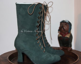 May include: A pair of dark green suede lace-up boots with a high heel. The boots have a vintage style with multiple eyelets and tan laces. The text "by Victoria Adriana" is visible on the side of the boot. A decorative dog figurine is in the background.