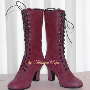 May include: A pair of burgundy knee-high boots with black laces and heels. The boots have a vintage style with a lace-up design and are made of a suede-like material. The text "by Adriana Popa" is visible on the side of the boots.