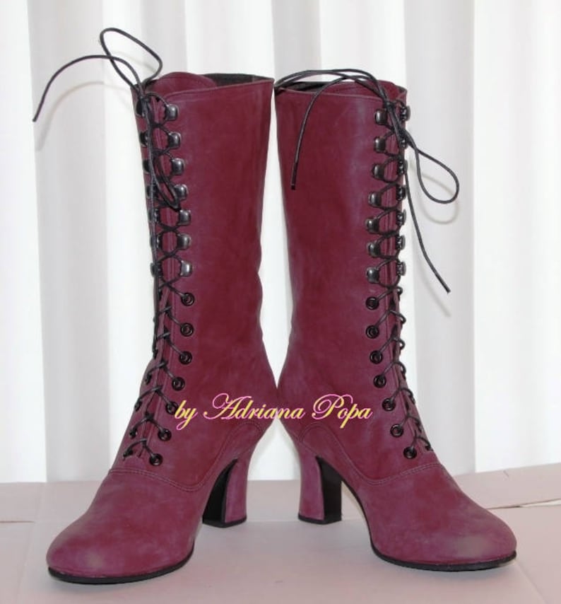 May include: A pair of burgundy knee-high boots with black laces and heels. The boots have a vintage style with a lace-up design and are made of a suede-like material. The text "by Adriana Popa" is visible on the side of the boots.