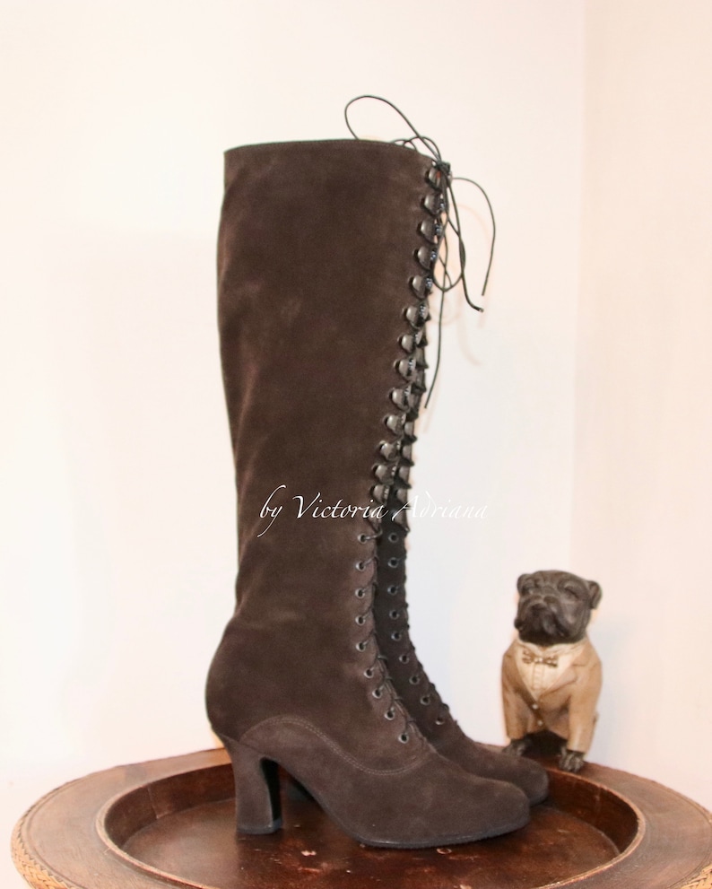 May include: Brown suede knee-high boots with a lace-up front and a block heel. The boots are on a wooden surface with a small dog figurine in the background.